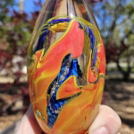Handheld oval memorial glass paperweight containing cremation ash, featuring flowing amber, gold, and deep blue glass layers with ribbon-like details, photographed outdoors in natural sunlight with a softly blurred background.