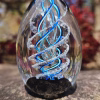 Outdoor front view of a spiral egg-shaped cremation glass keepsake featuring a clear glass form with vivid blue-aqua spiral detailing and visible cremation ashes suspended inside.