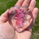 Clear glass eternity heart with soft pink flowing center design, handcrafted with cremation ash, resting in an open hand outdoors.
