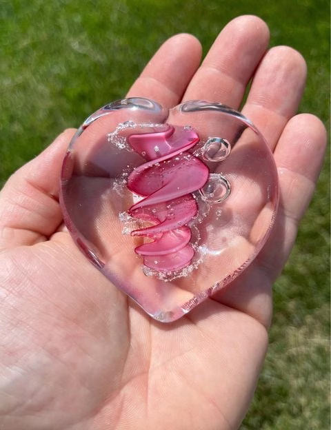 Clear glass eternity heart with soft pink flowing center design, handcrafted with cremation ash, resting in an open hand outdoors.