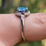 Side profile view of a sterling silver cremation ring featuring a square blue glass centerpiece, surrounded by 24 clear cubic zirconia accent stones set along the halo and band, shown worn on a finger outdoors.