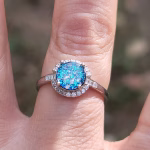 Top-down view of a sterling silver cremation ring featuring a round blue silver crinkle dichroic glass centerpiece, prong-set and surrounded by clear cubic zirconia accent stones, shown worn on a finger in natural sunlight.