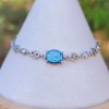 Centered front view of the Sands of Time Infinity Cremation Bracelet in blue silver crinkle, featuring an oval blue memorial glass stone with cremation ash, prong-set in sterling silver and connected by infinity-style links, displayed on a white jewelry stand with a softly blurred outdoor background.