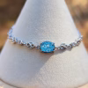 Front view of the Sands of Time Infinity Cremation Bracelet in blue silver crinkle, featuring an oval blue memorial glass stone with cremation ash, prong-set in sterling silver and connected by infinity-style links on a white jewelry display.