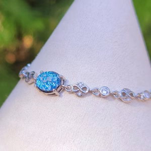 Angled side view of the Sands of Time Infinity Cremation Bracelet in blue silver crinkle, showing an oval blue memorial glass stone with cremation ash set in sterling silver, connected by infinity-style links and small accent stones, displayed on a white jewelry stand with a softly blurred green outdoor background.