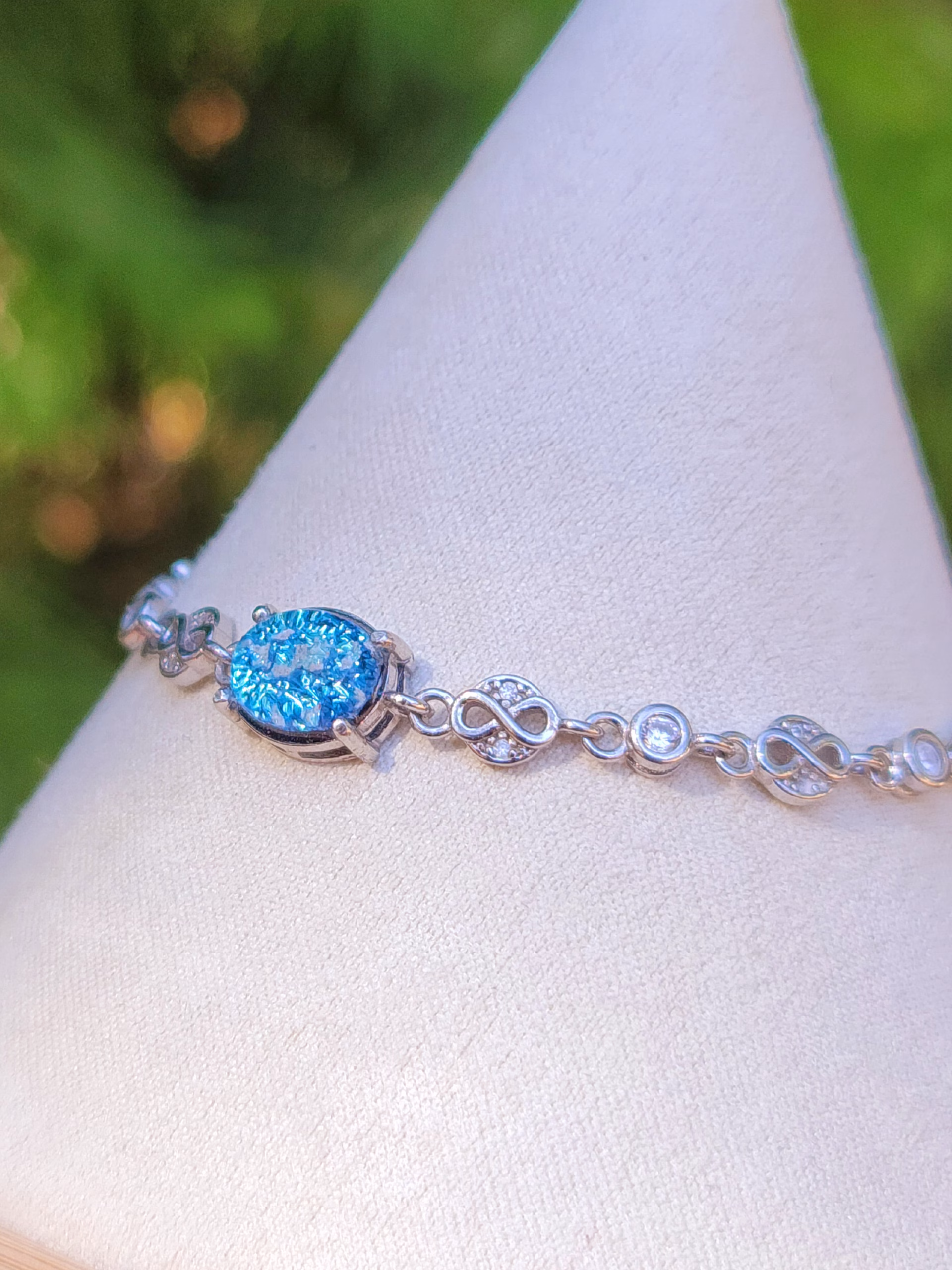 Angled side view of the Sands of Time Infinity Cremation Bracelet in blue silver crinkle, showing an oval blue memorial glass stone with cremation ash set in sterling silver, connected by infinity-style links and small accent stones, displayed on a white jewelry stand with a softly blurred green outdoor background.