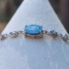 Front-facing close-up of the Sands of Time Infinity Cremation Bracelet showing a blue silver crinkle memorial glass stone set in sterling silver infinity links on a white jewelry display.