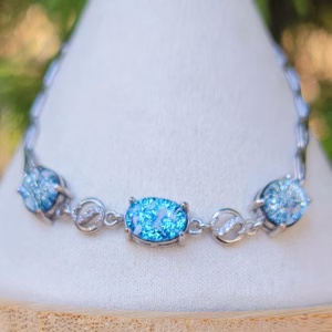 Front view of a sterling silver triple stone cremation bracelet displayed on a white jewelry stand, featuring three oval blue memorial glass stones with visible cremation ash, connected by polished silver links and subtle accent connectors.