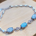 Sterling silver triple stone cremation bracelet laid in a circular shape on a light wooden surface, featuring three round blue memorial glass stones with visible cremation ash, connected by polished silver links and an adjustable clasp.