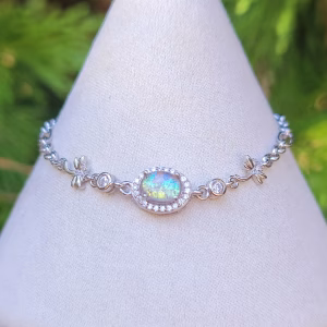 Front view of the dragonfly cremation bracelet featuring a clear rainbow memorial glass centerpiece set in sterling silver, accented with small dragonfly links and cubic zirconia details, displayed on a white jewelry stand with a soft green background.