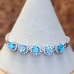 Front view of the Dichroic Halo Cremation Bracelet displayed on a white fabric jewelry stand, featuring five round blue dichroic memorial glass stones with visible cremation ash, each surrounded by clear cubic zirconia halos and set in sterling silver.