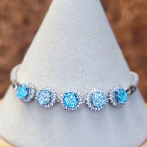Front view of the Dichroic Halo Cremation Bracelet displayed on a white fabric jewelry stand, featuring five round blue dichroic memorial glass stones with visible cremation ash, each surrounded by clear cubic zirconia halos and set in sterling silver.