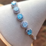Close-up angled view of the Dichroic Halo Cremation Bracelet featuring five round blue dichroic memorial glass stones, each surrounded by a halo of clear cubic zirconia, set in sterling silver and displayed on a white jewelry stand.