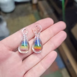 Rainbow Teardrop Cremation Pendant made with cremation ash, featuring layered rainbow colors encased in clear glass, shown resting in an open hand inside a glassblowing studio