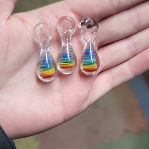 Rainbow Teardrop Cremation Pendants made with cremation ash, featuring layered rainbow colors encased in clear glass, shown as three pendants resting in an open hand