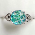 A sterling silver cremation ring featuring a round aqua crinkle memorial glass stone infused with visible cremation ash. The glass displays shimmering layers of blue, teal, and green, set in a four-prong mount with heart-shaped filigree accents on each side. The ring is photographed against a soft white background to highlight the vibrant colors and intricate details.