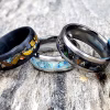 Cremation rings with colored stone inlay displayed on weathered wood, featuring multiple band styles in black and silver finishes with blue, green, copper, gold, and multicolor inlay options.