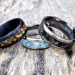 Cremation rings with colored stone inlay displayed on weathered wood, featuring multiple band styles in black and silver finishes with blue, green, copper, gold, and multicolor inlay options.