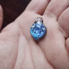 Handcrafted blue dichroic glass cremation pendant resting in the hand, featuring vibrant shimmering blue tones and scattered white flecks of encased cremation ashes beneath a smooth clear glass cap, shown close-up to highlight detail and scale