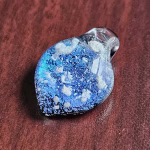 Handcrafted blue dichroic glass cremation pendant featuring shimmering metallic tones and scattered white flecks of encased cremation ashes beneath a clear glass cap, shown in a close-up view on a wooden surface to highlight color and texture