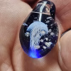 Handcrafted blue glass cremation pendant held between fingers, featuring a luminous white jellyfish suspended inside with shimmering white flecks of encased cremation ashes, shown in a bright close-up to highlight depth, color, and intricate glass artistry