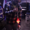Glass artist working in a dimly lit studio shaping a glowing molten glass form on a blowpipe with protective gloves and tools visible in the background