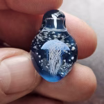 Handcrafted blue glass cremation pendant held between fingers, featuring a detailed white jellyfish suspended inside with swirling white flecks of encased cremation ashes, shown in a close-up view to highlight clarity, depth, and fine glasswork