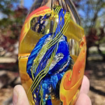 Handheld oval memorial glass paperweight with cremation ash suspended in layered blue, gold, and amber glass, featuring flowing ribbon details and reflective depth, photographed outdoors in natural light.