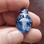 Handcrafted blue glass cremation pendant held between fingers, featuring an intricate white jellyfish suspended in swirling blue glass with shimmering white flecks of encased cremation ashes, shown close-up to highlight detail, depth, and craftsmanship