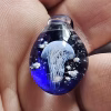 Handcrafted blue glass cremation pendant held between fingers, featuring a detailed white jellyfish suspended inside with swirling white flecks of encased cremation ashes, shown in a bright close-up to highlight depth, color, and fine glasswork