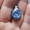 Handcrafted blue dichroic glass cremation pendant resting in the palm, featuring shimmering metallic blue tones and scattered white flecks of encased cremation ashes beneath a clear glass cap, shown close-up to highlight texture and size