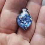 Handcrafted blue dichroic glass cremation pendant resting in the palm, featuring shimmering metallic blue tones and scattered white flecks of encased cremation ashes beneath a clear glass cap, shown close-up to highlight texture and size