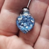 Handcrafted blue dichroic glass cremation pendant resting in the palm, featuring shimmering metallic blue tones and scattered white flecks of encased cremation ashes beneath a clear glass cap, shown close-up to highlight texture and size