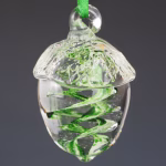 Hand-blown glass acorn memorial ornament featuring a clear acorn shape with swirling green coloration infused with cremation ash, hanging from a green ribbon against a dark background.