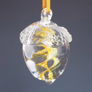 Hand-blown glass acorn memorial keepsake in transparent clear and gold tones, featuring a swirling gold design infused with cremation ash, suspended by a golden ribbon against a dark background.