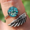 Angel Wing Cremation Ring worn on finger, featuring sterling silver wing band and round memorial glass centerpiece with teal and blue tones and cremation ash fused within the glass