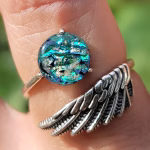 Angel Wing Cremation Ring worn on finger, featuring sterling silver wing band and round memorial glass centerpiece with teal and blue tones and cremation ash fused within the glass