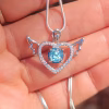 Angel heart cremation pendant in sterling silver with small angel wings on each side, a cubic zirconia–lined heart frame, and a round blue memorial stone at the center, photographed resting on a hand with the necklace chain visible.