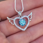 Angel heart cremation pendant in sterling silver with delicate wing details extending from each side of the heart, accented with cubic zirconia and a round blue memorial stone at the center, shown resting on a hand with the necklace visible.