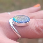A close-up side profile of the Bohemian Cremation Ring worn on a finger. The sterling silver band curves organically around an oval cremation stone with a rainbow-iridescent, textured surface containing ash. The angle highlights the stone’s low profile and the smooth wrap design of the band.