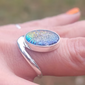 A close-up side profile of the Bohemian Cremation Ring worn on a finger. The sterling silver band curves organically around an oval cremation stone with a rainbow-iridescent, textured surface containing ash. The angle highlights the stone’s low profile and the smooth wrap design of the band.