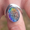 A sterling silver Bohemian-style cremation ring displayed on a finger, featuring an oval glass center with vibrant rainbow iridescence and visible cremation ash suspended within. The smooth silver band curves around the colorful focal stone for a bold, artistic look.