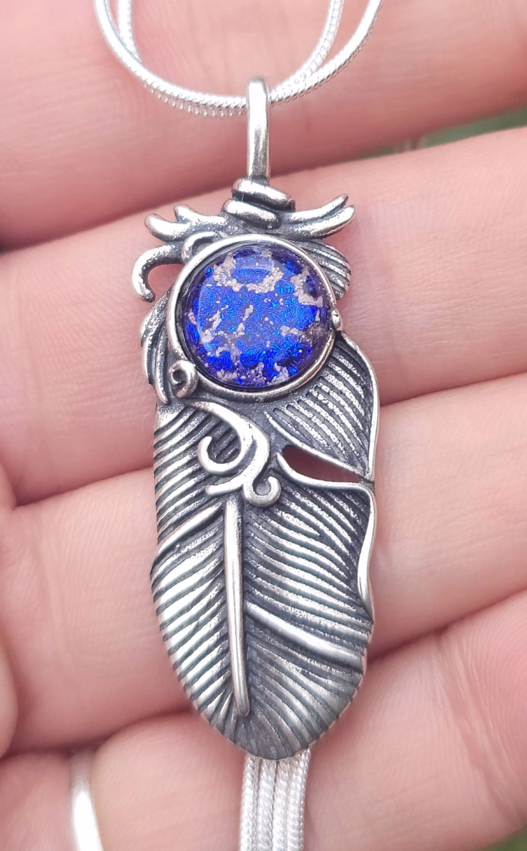 Close-up of the Boho Feather Cremation Pendant in sterling silver, featuring a feather memorial pendant with a blue cremation ash glass cabochon displayed against a hand for scale