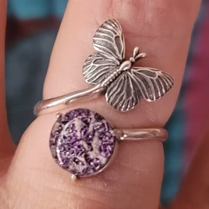 Close-up view of a sterling silver butterfly cremation ring featuring a detailed butterfly accent and a round purple glass centerpiece with cremation ash, shown worn on a finger against a softly blurred background.