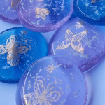 A close-up arrangement of blue and lavender glass cremation stones, each featuring gold butterfly designs and suspended cremation ash throughout the glass. The stones are smooth, rounded, and slightly translucent, showing subtle color variations and delicate detailing.
