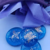 Blue glass cremation stones with gold butterfly designs and suspended cremation ash are arranged beneath a partially open purple drawstring pouch filled with lavender tissue paper. The stones display varied butterfly patterns and a smooth, polished finish.