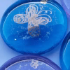 Close-up view of a blue glass cremation stone featuring delicate gold butterfly engraving and visible cremation ash suspended within the glass. Part of a purple butterfly stone is visible below it, showing variation in color and design.