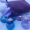 A lavender drawstring pouch with tissue paper peeking out sits beside a set of blue and purple glass cremation stones. Each stone contains visible cremation ash and is engraved with a delicate gold butterfly design. The stones are arranged in a loose spread on a light blue surface, showing their colors and details clearly.