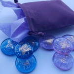 A lavender drawstring pouch with tissue paper peeking out sits beside a set of blue and purple glass cremation stones. Each stone contains visible cremation ash and is engraved with a delicate gold butterfly design. The stones are arranged in a loose spread on a light blue surface, showing their colors and details clearly.