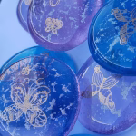 A collection of round glass cremation stones in shades of blue and lavender, each containing visible cremation ash and adorned with delicate gold butterfly engravings. The stones are arranged closely together, filling the frame, with light reflecting softly across their polished surfaces.